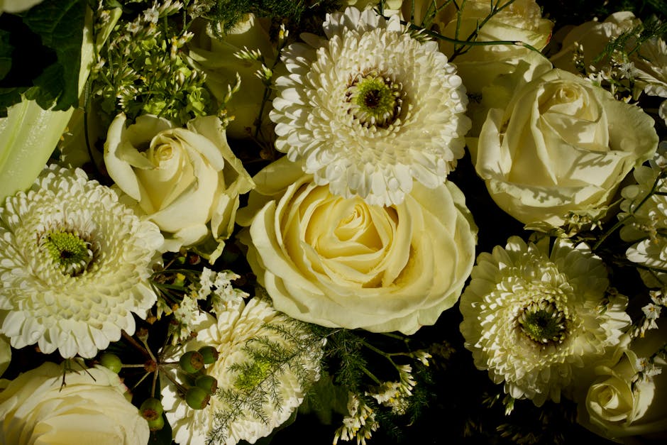 A close-up image of a floral arrangement featuring creamy white roses, large white Gerbera daisies, and smaller white flowers, accented with green foliage and delicate sprigs of fern. The roses displa