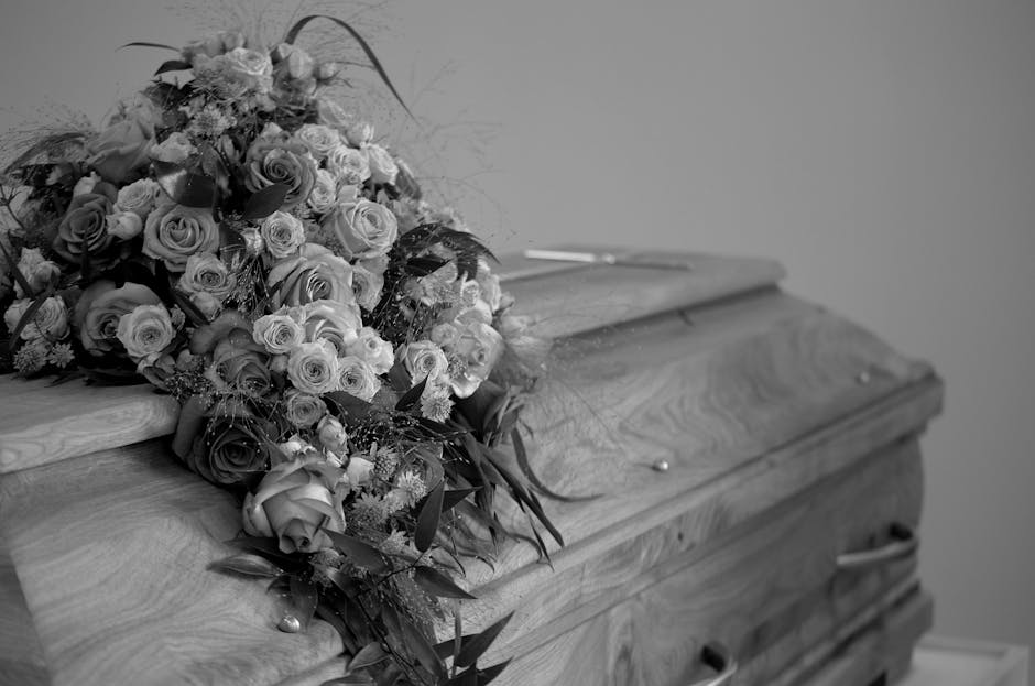 A large, carefully arranged floral tribute resting on top of a wooden casket. The bouquet features a mix of roses and other flowers in various sizes, with some fully bloomed and others as buds, surrou