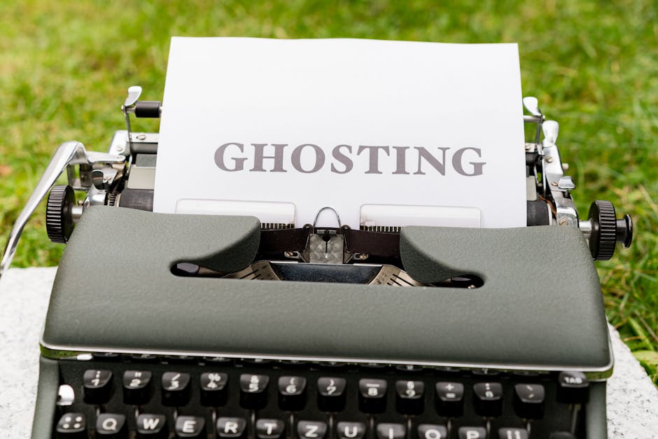 Close-up of a vintage typewriter outdoors on a white surface, with a sheet of paper inserted displaying the word 'GHOSTING' in bold, gray letters. The background shows green grass, indicating the scen