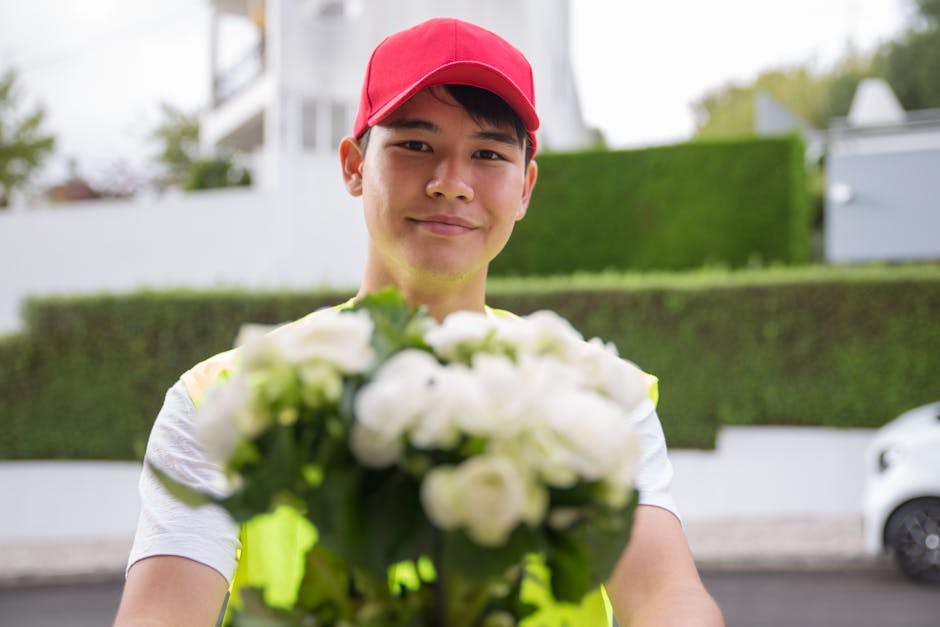 A delivery person wearing a red cap and a high-visibility vest is smiling and holding a bouquet of fresh white flowers, including roses and possibly peonies, wrapped in clear and pastel-colored wrappi