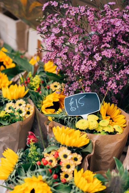A vibrant display of fresh floral bouquets arranged on a market stand, featuring large yellow sunflowers, small white daisy-like flowers, and sprigs of red and yellow accents, all wrapped in brown kra