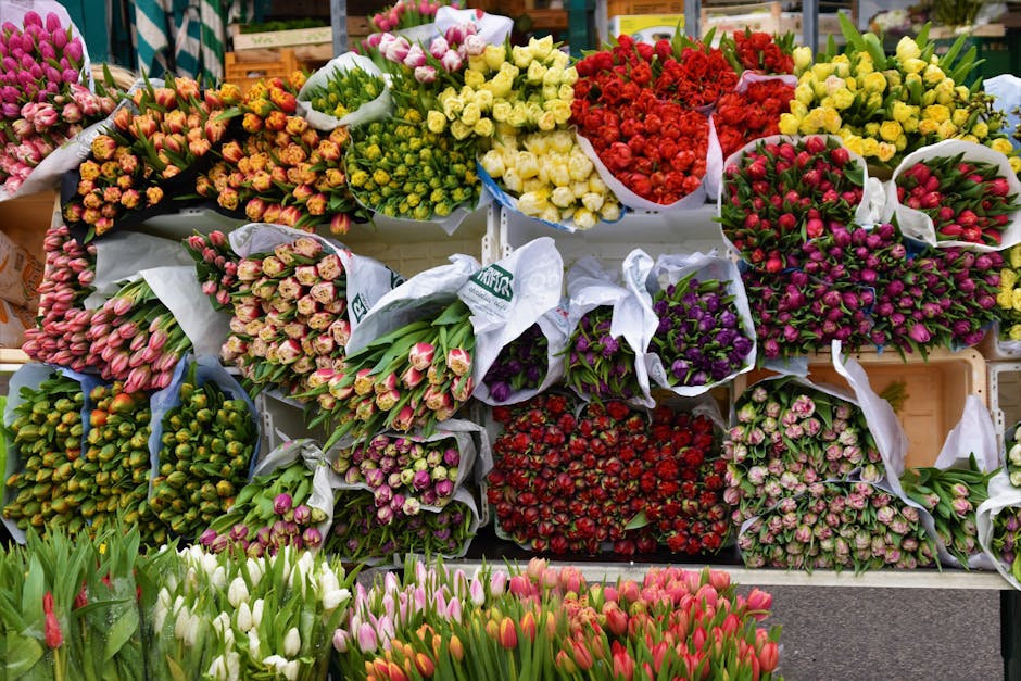 A vibrant display of fresh flowers arranged in bouquets at a florist shop, featuring tulips in pink, white, and red, alongside roses and other seasonal blooms. The bouquets are wrapped in white and pa