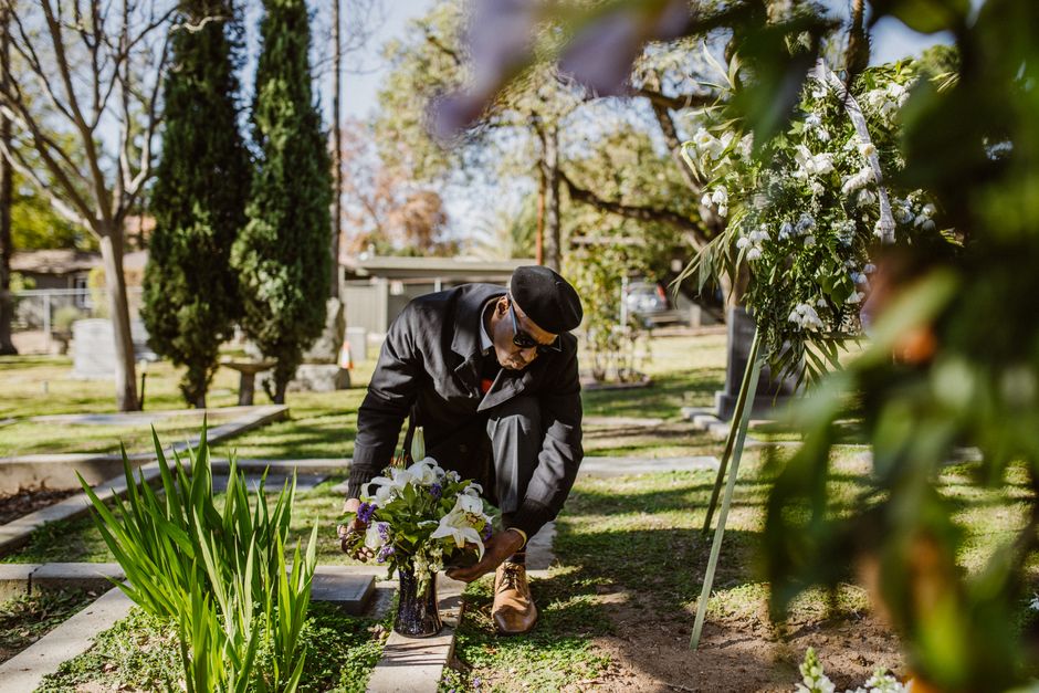 A man dressed in a dark suit, black hat, and sunglasses is kneeling in a cemetery, arranging a floral funeral tribute composed of white lilies, purple irises, and greenery in a black vase. He is caref