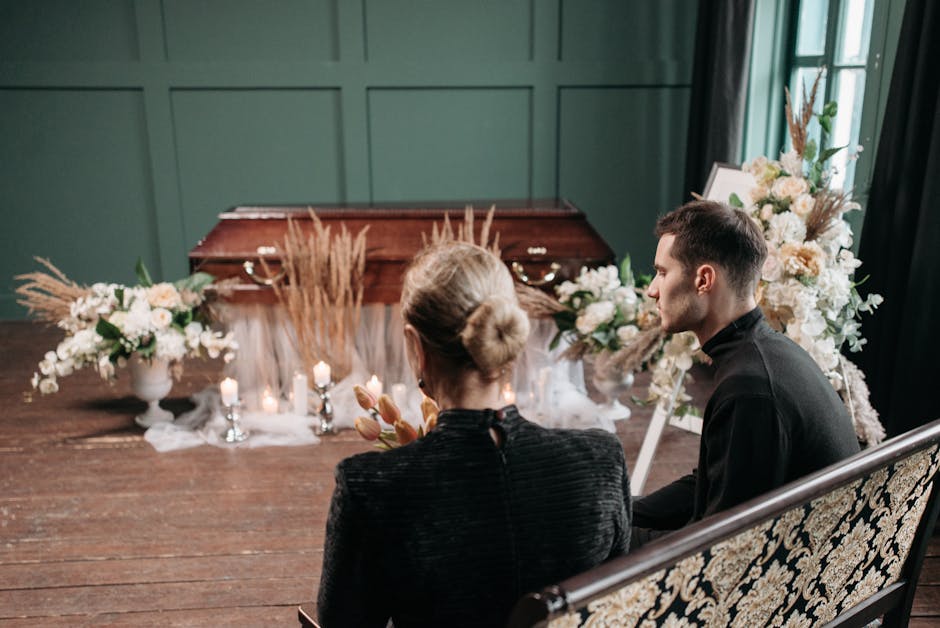 A close-up view of a floral arrangement at a funeral setting, featuring white roses, pale pink carnations, and dried beige pampas grass in elegant white vases. The flowers are arranged on a table drap